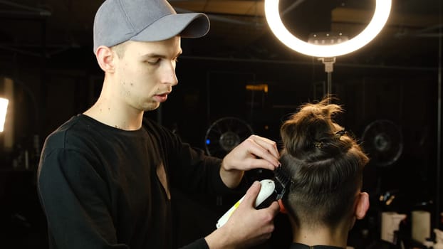 men's hairstyling and haircutting with hair clipper in a barber shop or hair salon. Hairdresser service in a modern barbershop in a dark key lightning with warm light back view.