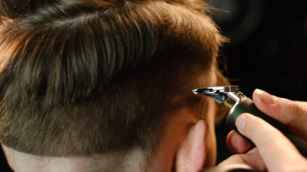 close up Shot of a handsome barber giving a haircut to his client using trimmer. Hairdresser service in a modern barbershop in a dark key lightning with warm light back view.
