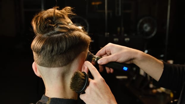 Shot of a handsome barber giving a haircut to his client using trimmer. Hairdresser service in a modern barbershop in a dark key lightning with warm light back view.