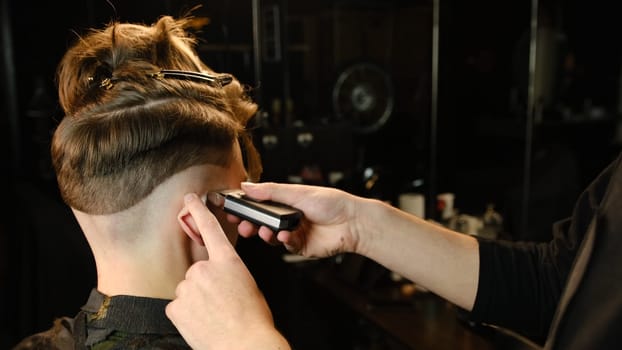 men's hairstyling and haircutting with barber shaving machine, hair cut for male. Hairdresser service with shaver in a modern barbershop in a dark key lightning with warm light back view.