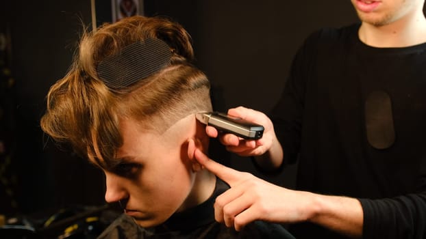men's hairstyling and haircutting with barber shaving machine, hair cut for male. Hairdresser service with shaver in a modern barbershop in a dark key lightning with warm light side view.