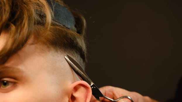 Close up Barber master hairdresser does hairstyle and style with scissors and comb. Concept Barbershop. side view in dark key lightning with warm light.