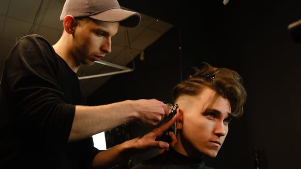 men's hairstyling and haircutting with hair clipper in a barber shop or hair salon. Hairdresser service in a modern barbershop in a dark key lightning with warm light side view.