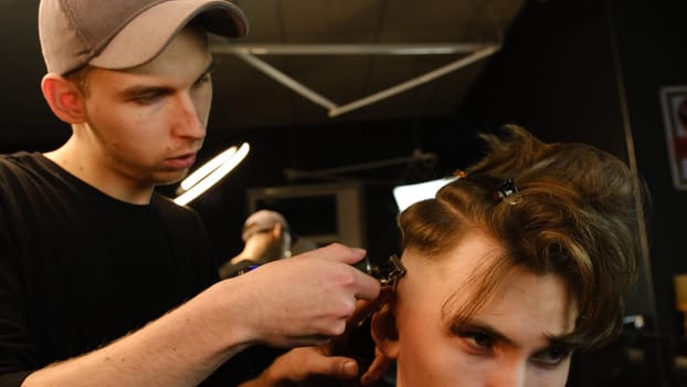 Shot of a handsome barber giving a haircut to his client using trimmer. Hairdresser service in a modern barbershop in a dark key lightning with warm light side view.