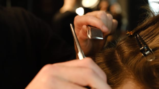 Close up Barber use Thinning scissors and metal comb on brown wavy hair of young man. Hairdresser service in a modern barbershop in a dark key lightning with warm light side view.