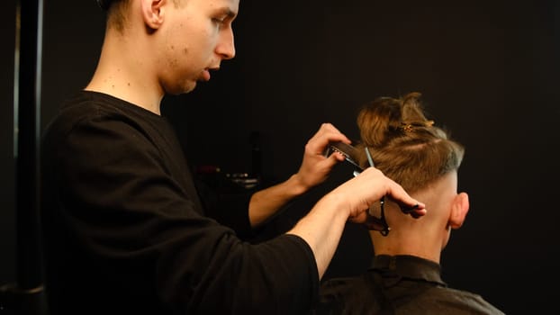 Barber use Thinning scissors and metal comb on brown wavy hair of young man. Hairdresser service in a modern barbershop in a dark key lightning with warm light back view.