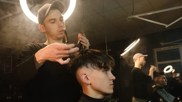 Barber master hairdresser does hairstyle and style with scissors and comb. Concept Barbershop. side view in dark key lightning with warm light and smoke.