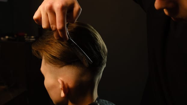 Barber use Thinning scissors and metal comb on brown wavy hair of young man. Hairdresser service in a modern barbershop in a dark key lightning with warm light.