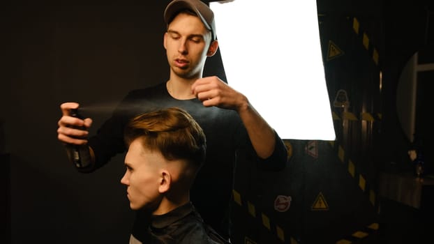 Barber makes hair styling with hair spray after haircut at the barber shop. Young handsome Caucasian man getting a haircut in a modern hairsalon from professional hairdresser.