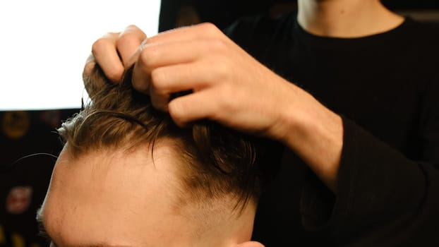 Barber shop. Man in barber's chair, hairdresser styling his hair with hands in dark key lightning. Hairdresser for man.
