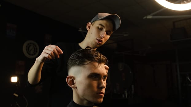 Barber shop. Man in barber's chair, hairdresser styling his hair with hands in dark key lightning. Hairdresser for man.