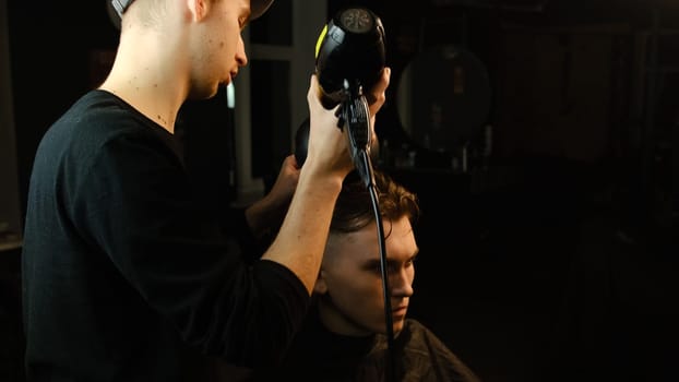 Young barber drying hair of customer with hair dryer in barber shop in a dark key lightning with warm light and correct hair with hand. Hairdresser for man.