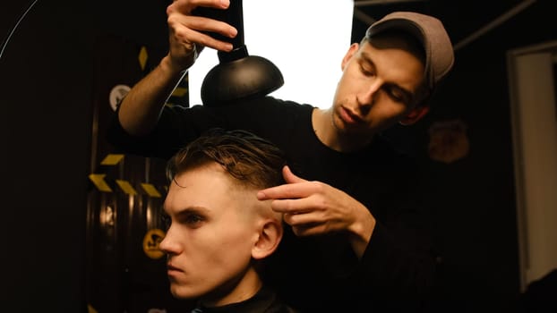Young barber drying hair of customer with hair dryer in barber shop in a dark key lightning with warm light and correct hair with hand. Hairdresser for man.