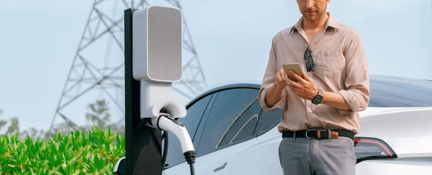 Man pay for electricity with smartphone while recharge EV car battery at charging station connected to electrical lower grid tower for eco friendly car utilization. Panorama Expedient