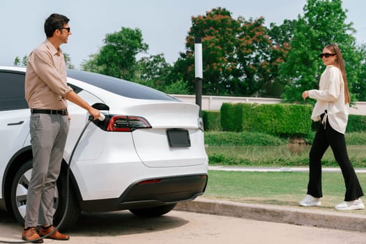 Young couple recharge electric car's battery from charging station in outdoor green city park in springtime. Rechargeable EV car for sustainable environmental friendly urban travel lifestyle.Expedient