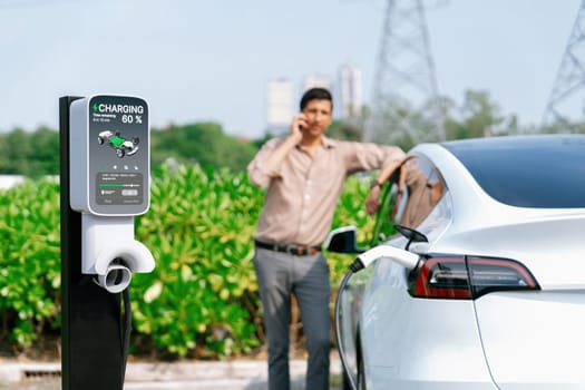 Man talking on the phone while recharge EV car battery at charging station connected to power grid tower electrical as electrical industry for eco friendly car utilization.Expedient