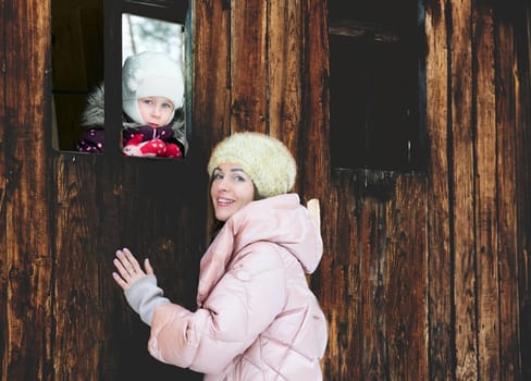 a woman in relation to her child or children. Pretty mother with a child in the window of a wooden toy house in a winter park.