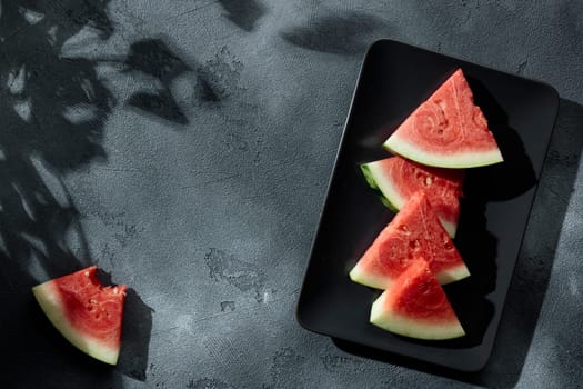 Slices of juicy watermelon on the black plate on the concrete background with summer sun shadows. Copy space. Top view.