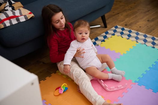 Adorable girl lovely sister holding supporting her younger brother, a cute baby boy trying to do his first steps, sitting on multi colored puzzle carpet at home. Children. Growth. Family relationships