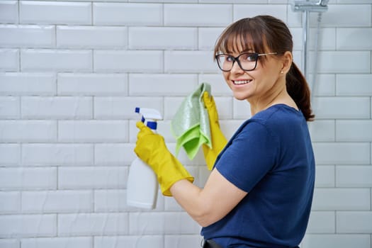 Woman with detergent spray, rag cleaning in bathroom, washing white wall tiles, smiling female looking at camera. Routine house cleaning, home hygiene, housecleaning service, housekeeping, housework