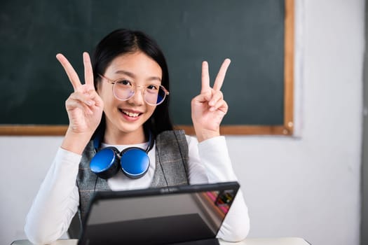 A young girl wearing glasses and a blue vest is sitting at a desk with a laptop in front of her. She is smiling and holding up her hands in a peace sign