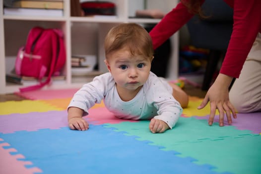 Selective focus on Caucasian beautiful adorable baby boy crawling on colorful puzzle carpet with the help of his young loving caring sister. Children growth and development. Babyhood. Infancy. Family