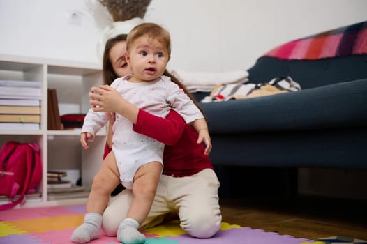 Adorable Caucasian little child girl, loving caring sister holding and supporting her little brother, an adorable baby boy trying to his first steps on a colorful puzzle carpet. Family relationships.