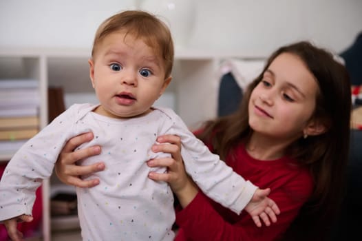 Authentic portrait of beautiful Caucasian kids. Lovely baby boy 6-9 months old and his loving caring sister 7 years old plying together at home