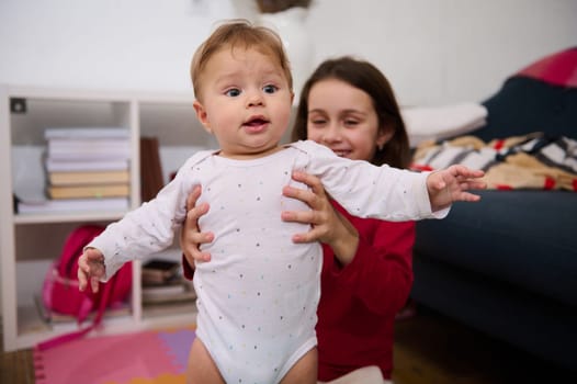 Smiling baby boy in the hands of his loving caring sister. Cute child girl supporting her brother doing first steps. Infancy and babyhood. Happy family relationships. Support. Spending time together