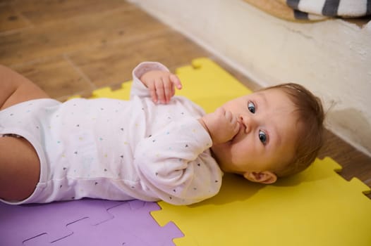 European cute lovely baby boy 6-8 months old lying on multicolored puzzle carpet, suckling his finger, looking at camera with curiosity. Beautiful babies. Infancy Babyhood. Kids growth and development