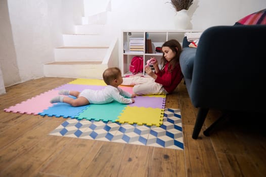 Side portrait of adorable little child girl, loving caring sister playing with her baby boy brother on a colorful puzzle carpet at cozy home interior. Happy family and World Children's Day concept
