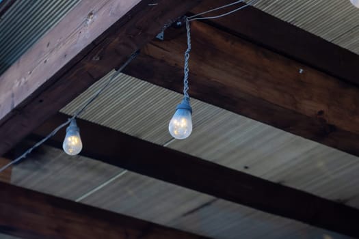 A string of lights is suspended from a sturdy wooden beam, adding a warm ambiance to the shaded area below. The hardwood structure is accented with wood stain, creating a cozy atmosphere by the water