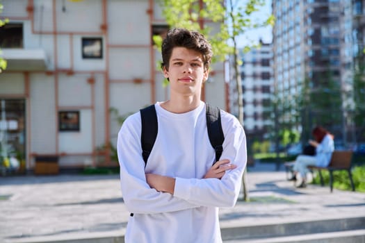 Portrait of confident smiling college student guy, young male with crossed arms with backpack looking at camera outdoor near educational building. Education, training, 19,20 years age youth concept