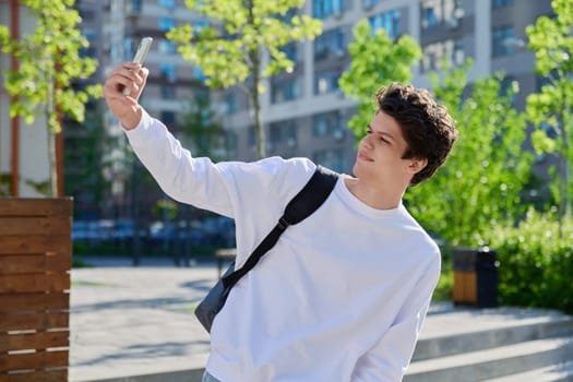 Young guy student with smartphone in his hands taking selfie outdoor portrait, male university college student. Youth, urban style, 19,20 years man, lifestyle concept