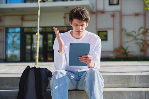 Young handsome guy college student using digital tablet outdoor, talking on video call chat conference, educational building background. Education, technology, training, 19,20 years age youth concept