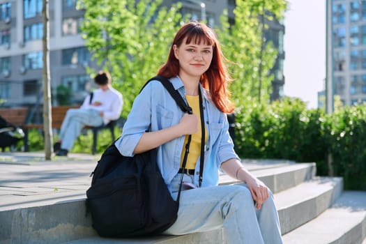 Portrait of young beautiful female college student outdoor, smiling red-haired girl 19, 20 years old with backpack looking at camera, educational building background. Education training youth concept