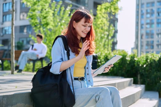 Young attractive female college student using digital tablet outdoor, talking on video call chat conference, educational building background. Education, technology, training, 19,20 years age youth concept