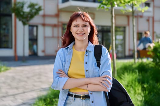 Portrait of young beautiful female college student outdoor, smiling red-haired confident girl with crossed arms backpack looking at camera, educational building background. Education training youth