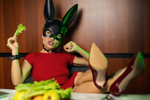 A beautiful girl in a bdsm rabbit mask and a bright red dress eats lettuce leaves promoting a healthy lifestyle and vegetarianism