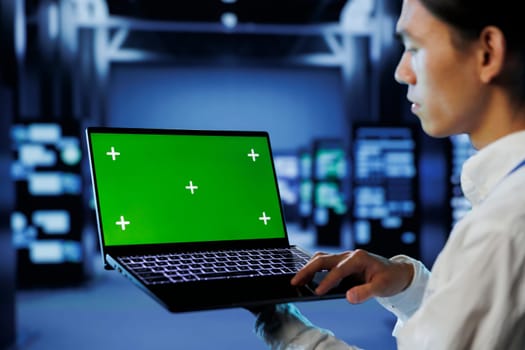 IT consultant with green screen laptop between server hub clusters providing processing resources for businesses worldwide. Worker uses mockup device to fix data center mainframes