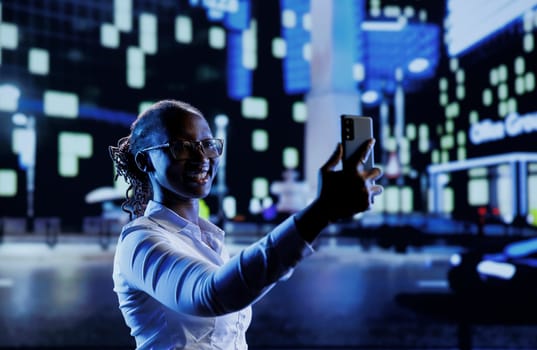 BIPOC woman walking around city at night, using mobile phone to take selfie. Citizen using smartphone to take pictures while strolling outside on empty streets illuminated by lamps