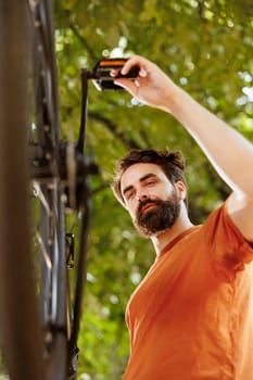 Detailed outside image shows youthful healthy caucasian man inspecting and maintaining bicycle components. Dedicated male cyclist outdoors repairing bike crank arm and pedal for summer cycling.