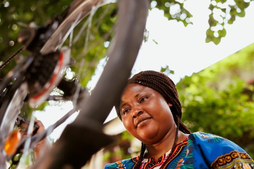 Young sports-loving black woman examining bicycle tire in home yard for damage to repair with tools. Energetic african american female fixing broken bike chain and wheel outside.