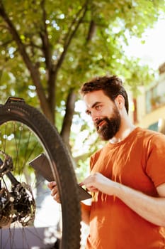 Enthusiastic healthy male cyclist checking tire as yearly bike rubber maintenance routine with digital tablet. Caucausian man using smart smart device to look for solutions of damaged bicycle tire.