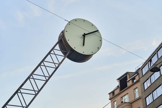 A clock hangs from a metal pole against the sky, in front of a building. The circular clock face contrasts with the urban design of the tower and windows