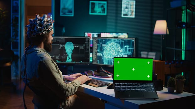 Engineer with EEG headset on programming brain transfer into computer virtual world next to green screen laptop. Transhumanist using neuroscience to gain digital soul, mockup device, camera B