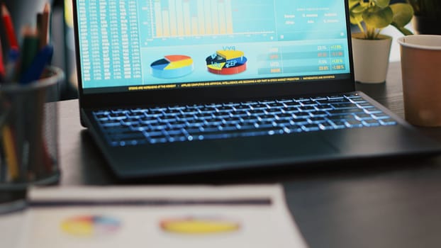 Financial report on desk next to laptop showing marketing statistics in empty office, close up shot. Company files containing business pie charts near notebook left open in workspace