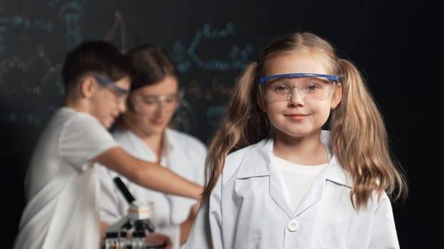 Smart girl looking at camera while teacher and student doing experiment behind. Pretty child looking at camera while boy looking under microscope at black board written chemical theory. Erudition.