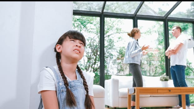 Stressed and unhappy young girl huddle in corner, cover her ears blocking sound of her parent arguing in background. Domestic violence at home and traumatic childhood develop to depression. Synchronos