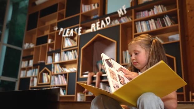 Young smart caucasian girl picking reading a book while sitting at library. Clever child learning, studying, open a books at library. Attractive kid turning page with blurring background. Erudition.